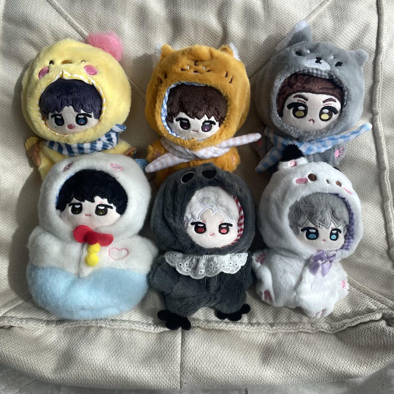 May include: Six plush dolls dressed in various animal-themed hooded outfits. The dolls feature embroidered facial details, including large eyes and hair. The outfits include a yellow chick, orange fox, gray wolf, white seal, dark gray bird, and white cat.