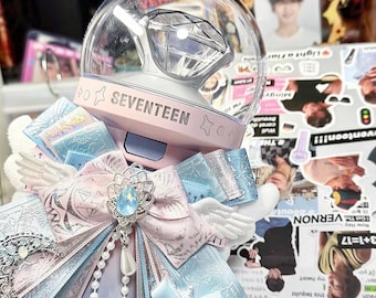 May include: A light pink and white SEVENTEEN lightstick featuring a clear dome with a diamond-shaped insert. The handle is decorated with layered blue and pink patterned ribbons, white angel wings, a silver moon charm, and a blue gem.