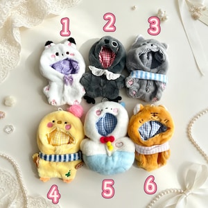 May include: Six plush animal-themed doll outfits with hoods, numbered 1 through 6. The collection includes a white cat, a dark gray bird, a light gray cat, a yellow chick, a white and blue character, and an orange tiger, all with fabric linings.