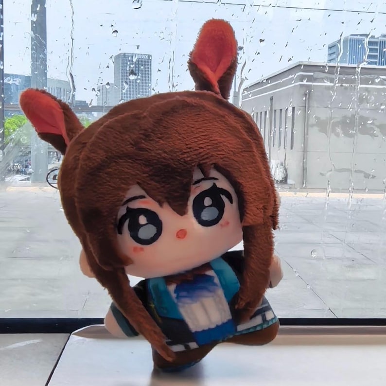 May include: A plush doll with brown hair and red ears, wearing a blue and white outfit. The doll has large, expressive eyes and is positioned in front of a window with a city view.