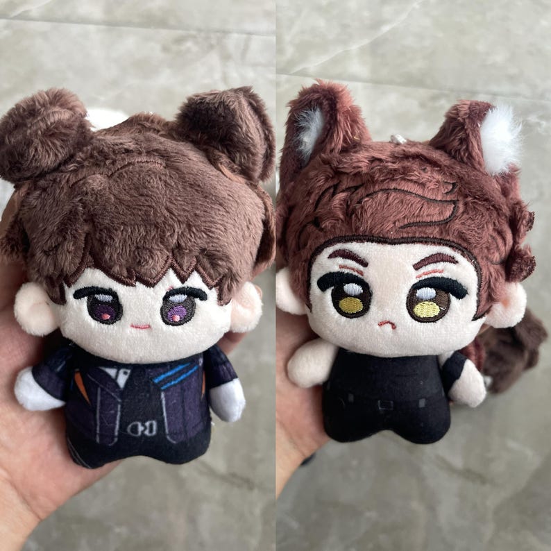 May include: Two plush dolls with brown hair and embroidered facial features. The left doll has round ears and a dark jacket with blue accents. The right doll features pointed animal ears with white tips, a brown tail, and a plain black outfit.