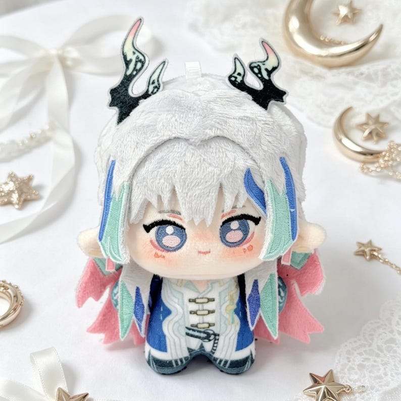 May include: A plush doll with white hair and blue and green accents. The doll has large blue eyes and is wearing a blue and white outfit with pink accents. The doll has black and white decorative horns.