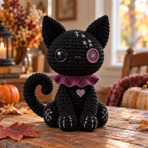 Puede incluir: Un gato de peluche tejido a ganchillo en color negro con un ojo de botón negro brillante grande y otro de botón morado más pequeño. Tiene un cuello con volantes morado, un pequeño corazón bordado en el pecho y detalles cosidos en blanco.