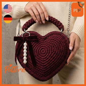 May include: A burgundy heart-shaped crochet bag with a braided handle and silver chain strap. A burgundy velvet bow and a string of white pearls adorn the front. The bag is held by a person wearing a cream-colored sweater. The word "Pattern" is visible.