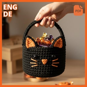 May include: A black crochet cat-shaped basket with orange ears and nose, filled with wrapped candies. The basket has a handle and is held by a hand. The basket is a Halloween-themed item.