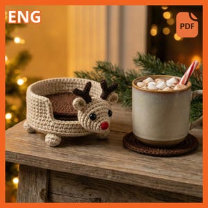 May include: A crocheted reindeer bed with a brown interior and a reindeer head on the front. A mug of hot chocolate with marshmallows and a candy cane sits on a coaster next to the bed. The bed is on a wooden surface.