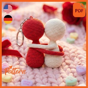 Peut inclure: Un porte-clés représentant un couple crocheté, l'un rouge et l'autre blanc, s'embrassant. Le personnage rouge a un anneau de porte-clés argenté. Le fond comprend une surface tricotée rose, des bonbons en forme de cœur et un ruban rouge. Le mot "Pattern" est visible.