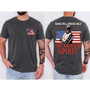 Puede incluir: Camiseta gris oscuro con un diseño patriótico. La parte delantera presenta una bandera estadounidense estilizada y el texto "Dale". La parte trasera muestra una foto de un piloto de carreras, la bandera estadounidense y las palabras "RAISE HELL, PRAISE DALE" y "THE AMERICAN SPIRIT."
