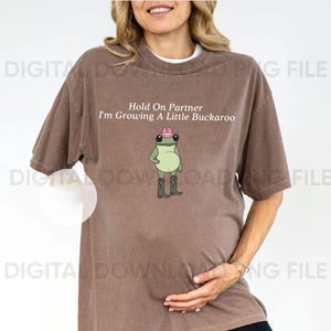 May include: Brown t-shirt with the text "Hold On Partner I'm Growing A Little Buckaroo" and a cartoon frog wearing a pink cowboy hat and boots. The shirt has a crew neck and short sleeves.