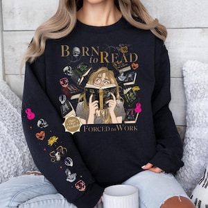 May include: Black sweatshirt with the text "Born to Read Forced to Work" and a graphic of a person reading surrounded by books. The left sleeve features colorful illustrations of skulls, flowers, and musical instruments. A cozy and stylish garment for book lovers.
