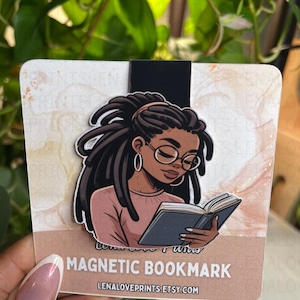 May include: A magnetic bookmark featuring an illustration of a Black woman with locs, wearing glasses, and reading a book. The bookmark has a black magnetic strip and the text "MAGNETIC BOOKMARK" and "LENALOVEPRINTS.ETSY.COM". The background is a watercolor design.