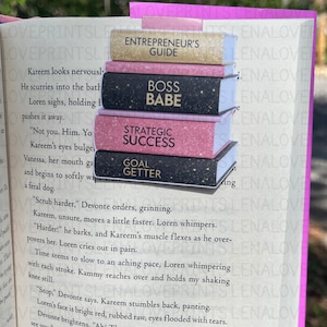 May include: A stack of four book-shaped bookmarks with motivational phrases. The top bookmark reads "ENTREPRENEUR'S GUIDE" in gold, followed by "BOSS BABE" in black, "STRATEGIC SUCCESS" in pink, and "GOAL GETTER" in black. The bookmarks are placed on the pages of a book.