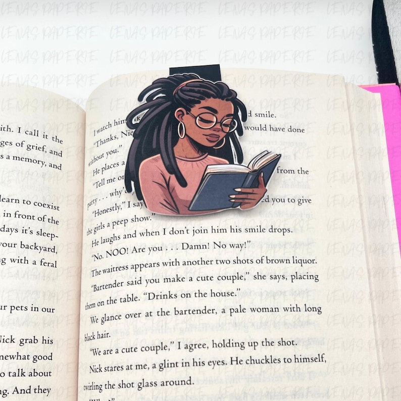 May include: A bookmark featuring an illustration of a Black woman with locs, wearing glasses, and reading a book. The bookmark is placed on the pages of a book. The woman is wearing a pink shirt.