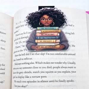 Pode incluir: Marcador de livros com uma mulher negra de cabelo encaracolado e &oacute;culos, segurando uma pilha de livros. Os livros t&ecirc;m t&iacute;tulos como "Be Rested" e "Be At Peace". Um acess&oacute;rio perfeito para os amantes da leitura.