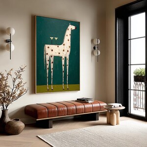 May include: An abstract painting of a giraffe with brown spots and two martini glasses on its back, set against a teal background. The artwork is framed in gold and hangs above a brown leather bench in a modern living room.
