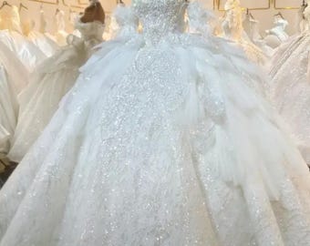 Puede incluir: Un vestido de novia blanco sin tirantes con un corpiño muy adornado y una falda amplia y en capas. El vestido está cubierto de lentejuelas brillantes y detalles intrincados, creando un look glamuroso y elegante. Otros vestidos blancos son visibles en el fondo.