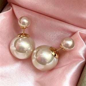 May include: A pair of double-sided earrings featuring two spherical white pearls of different sizes connected by a gold-toned metal post. The larger pearl sits at the base, while the smaller pearl serves as the front stud component of the jewelry.