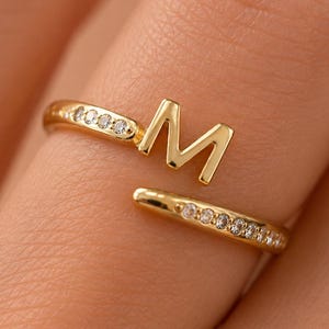 May include: A gold-toned open ring featuring a capital letter M as a central focal point. The band is split into two ends, each embellished with a row of small, clear, sparkling stones set along the metal surface, creating a delicate appearance.