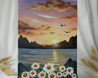 May include: A small rectangular painting on a wooden easel shows a sunset over a lake. The sky fades from purple to orange, with silhouetted mountains and flying birds. White daisies with yellow centres are in the foreground.
