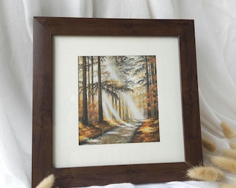 May include: A framed landscape painting of a forest scene with sunlight filtering through the trees. The artwork shows a path, with warm autumn colours and a dark brown wooden frame.