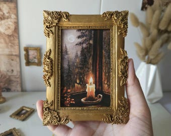 May include: A small rectangular painting in an ornate gold frame featuring a lit candle on a windowsill overlooking a dark, rainy forest scene with a full moon in the sky. The frame has intricate decorative carvings at each corner and along sides.