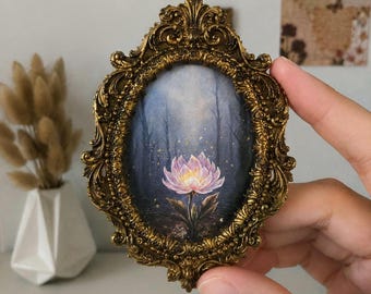 May include: An oval-shaped picture frame with an ornate, antique gold finish featuring intricate scrollwork and leaf motifs. The frame contains a painting of a single pink lotus flower centred against a dark, misty forest background with gold specks.