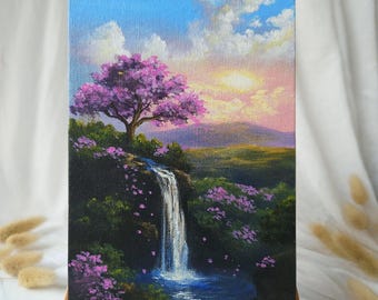 Puede incluir: Una pintura de paisaje sobre lienzo representa una cascada que cae en un arroyo azul. Un árbol con flores moradas se alza sobre una colina, bajo un cielo con nubes y un atardecer. La pintura se exhibe en un pequeño caballete de madera.