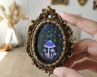 May include: A small oval frame with an ornate, metallic gold finish containing a dark painting of glowing blue and purple mushrooms in a forest setting. The frame features intricate scrollwork and decorative detailing along the entire border.