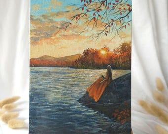 May include: An original oil painting on canvas showing a tranquil lakeside sunset. A figure in an orange dress sits on a rock, looking at the water. The sky shifts from blue to orange, with trees and mountains in the background. The painting is displayed on a small wooden easel.