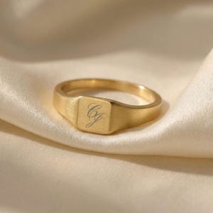 Pode incluir: Um anel de sinete dourado com uma face quadrada e um acabamento escovado. O anel apresenta as iniciais gravadas "Cg" em uma escrita delicada. O anel é exibido em um tecido de cetim creme, destacando sua aparência polida.