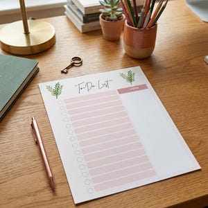 Op de afbeelding: Een To-Do List notitieblok met een roze en wit ontwerp, met een lijst met selectievakjes en een notitiesectie. Het notitieblok ligt op een houten bureau, naast een roségouden pen, een sleutel en een lamp.