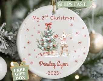 May include: A white ceramic Christmas ornament with a festive design. It features a Christmas tree, a bunny, and the text "My 2nd Christmas" and "Presley Lynn -2025-". Red snowflakes and a bow add to the holiday theme.
