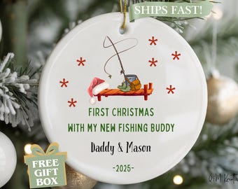 May include: White Christmas ornament with a fishing theme. It features a Santa hat, fishing rod, and a small dock. Text reads "First Christmas with my new fishing buddy" and personalized names and year.