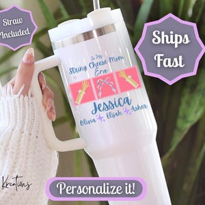 May include: White insulated tumbler with a handle, personalized with the name "Jessica" and the text "In My String Cheese Mom Era." Includes a straw. The design features pink and blue accents. The tumbler is held by a person wearing a white sweater.