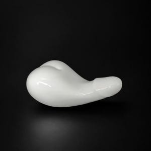 May include: A smooth, white, abstract sculpture with a rounded form and a protruding, cylindrical shape. The object is set against a black background, highlighting its glossy surface and unique design.