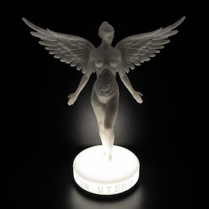 May include: A white sculpture of a winged figure with outstretched arms, standing on a glowing circular base. The sculpture has detailed wings and visible internal organs. The base reads "IN UTERO" in white lettering. The background is black.