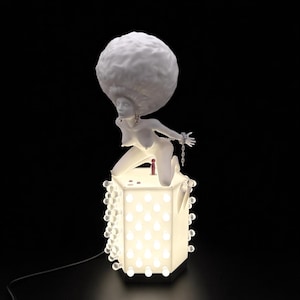 May include: A white sculpture of a woman with a large afro kneeling on a glowing cube-shaped lamp. The woman's wrists are chained. The lamp has a series of small, round lights around the edges and inside the cube. A black power cord extends from the lamp.