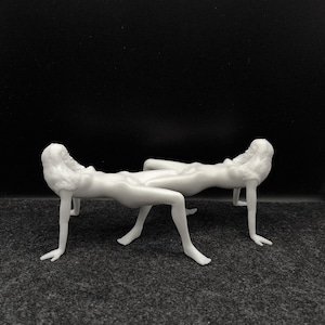 May include: Two white, stylized human figure sculptures in a reclining pose, supported by their arms and legs. The figures are positioned side-by-side against a dark background. The sculptures have detailed facial features and flowing hair.