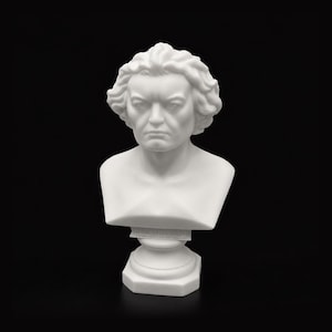 May include: A white matte bust sculpture of Ludwig van Beethoven featuring his signature wavy hair, a stern facial expression, and a focused gaze. The figure is mounted on a pedestal base with the name BEETHOVEN embossed on the front section.