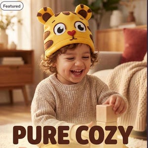 Puede incluir: Un gorro de forro polar amarillo con diseño de cara de tigre, rayas marrones, dos orejas redondeadas, grandes ojos blancos con pupilas negras y una pequeña nariz roja en forma de corazón. El texto PURE COZY aparece en la parte inferior.