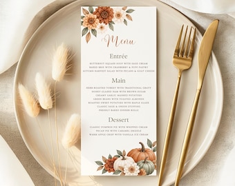 May include: A rectangular paper menu with floral and pumpkin illustrations, featuring the text Menu, Entrée, Main, and Dessert, placed on a round plate with a gold fork and knife, accompanied by three dried beige grass stems.