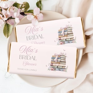 Könnte beinhalten: Zwei rosa Schokoriegelverpackungen mit Goldkanten, mit dem Text "Mia's BRIDAL Shower" und einem Stapel Bücher mit rosa Schleife und floralen Akzenten. Eine Verpackung sagt auch "THANKS FOR COMING!"