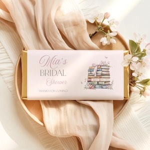 Puede incluir: Una envoltura rectangular para barra de chocolate en rosa pálido con extremos de papel dorado. El texto dice Mia's BRIDAL Shower y THANKS FOR COMING! A la derecha hay una ilustración de una pila de libros con un lazo y flores.