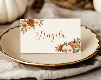 May include: A rectangular place card with a cream background featuring the name Angela in elegant brown script. The design includes watercolour illustrations of orange and white pumpkins, brown and white flowers, and green leaves.