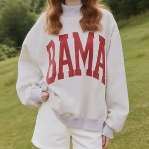 May include: A light gray sweatshirt with the word "BAMA" in large, red, block letters. The sweatshirt has gray trim at the collar, cuffs, and hem. The person is wearing white shorts. The background is a green, grassy area.