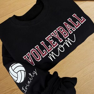 May include: Black sweatshirt with the words "VOLLEYBALL mom" in maroon and white lettering. The sleeve features a white volleyball graphic and the name "trinity" in script. The sweatshirt is made of a soft material.