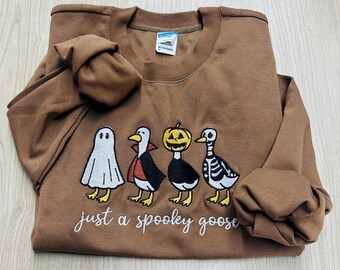May include: A brown sweatshirt featuring a Halloween-themed design. The design includes a ghost, a vampire duck, a pumpkin duck, and a skeleton duck. The phrase "just a spooky goose" is embroidered below the characters.