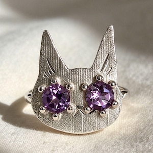 May include: A silver-toned ring featuring a cat-shaped face with a textured, brushed metal finish. The cat's eyes are represented by two round, faceted purple gemstones set securely into the metal. The design includes pointed ears and a small, engraved V-shaped nose detail.