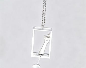 May include: A silver-toned pendant necklace featuring a rectangular frame containing a sculpted arm and hand reaching upward. A small, circular silver-toned charm hangs from the bottom of the frame, suspended by a delicate silver-toned chain.