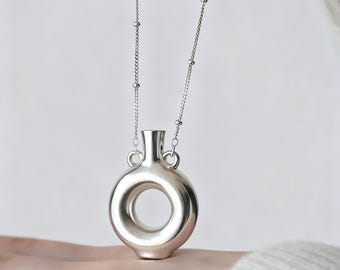 May include: A silver-toned pendant shaped like a small, hollow ring with a narrow neck at the top, suspended from a delicate silver chain featuring small, evenly spaced spherical beads along its length, creating a polished metallic appearance.